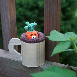 May include: A wooden mug with a brown crocheted rim and a crocheted carrot with a green top inside. The mug is sitting on a wooden surface.