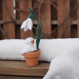 May include: A felt snowman holding a small potted plant with a white flower. The plant is in a brown pot and the snowman is wearing a white hat.