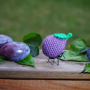 May include: Three purple plums and a crocheted purple plum with green leaves on a wooden surface with green leaves.