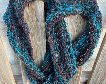 May include: A brown and turquoise knitted scarf with a loose, open weave. The scarf is draped over a wooden fence.