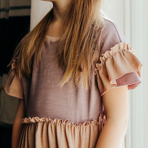 May include: A two-tone dress in shades of mauve and peach. The dress features ruffled sleeves and a ruffled waistline. Two large patch pockets are sewn onto the front of the dress. The dress is made of a soft, lightweight fabric.