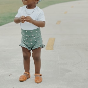 May include: A young child wearing a white t-shirt, green floral print shorts, and brown sandals. The child is wearing a green headband with a bow. The child is standing on a paved path.