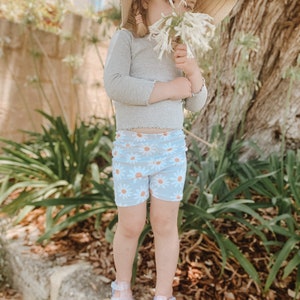 May include: A young girl wearing a grey long-sleeved shirt, blue daisy print shorts, and clear jelly sandals. She is holding a bouquet of white flowers and wearing a straw hat with a brown ribbon.