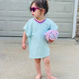 May include: A young girl wearing a light blue t-shirt dress with a purple trim, pink sunglasses, and a pink and blue patterned towel.
