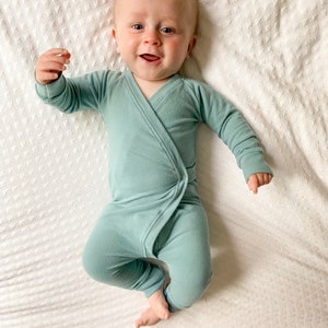 Könnte beinhalten: Ein Baby trägt einen hellblauen, langärmeligen Strampler mit Wickelverschluss. Das Baby liegt auf einer weißen Decke und lächelt.