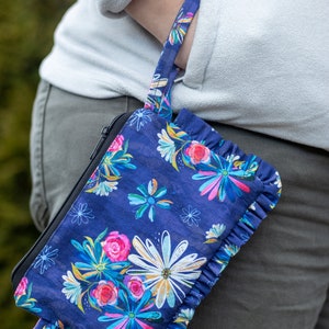May include: A blue fabric pouch with a floral print and a ruffled edge. The pouch has a black zipper and a black strap.