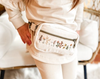 May include: A cream-colored fanny pack with a zipper closure and embroidered floral designs. The fanny pack is worn around the waist of a child wearing a cream-colored sweater and pants.