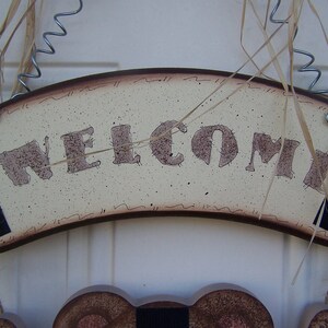 May include: A wooden welcome sign with a brown border and a cream background. The word "WELCOME" is written in brown letters with a distressed look. The sign is hanging from a wire and twine.
