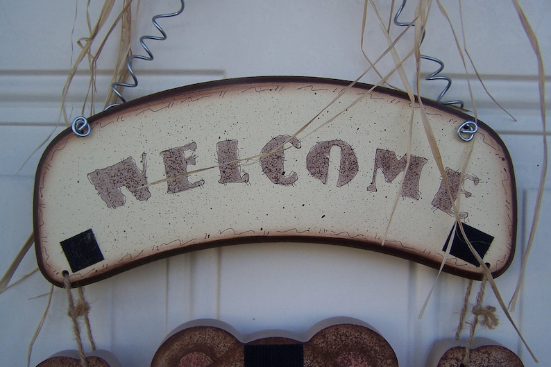 May include: A wooden welcome sign with a brown border and a cream background. The word "WELCOME" is written in brown letters with a distressed look. The sign is hanging from a wire and twine.