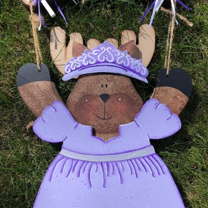 May include: A wooden moose wearing a purple dress with a silver belt and a purple crown. The moose is holding up its arms and has a happy expression. The moose is hanging from a wooden sign that says "WELCOME".