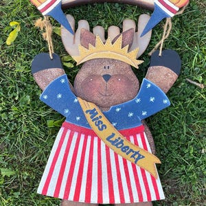 May include: A wooden moose decoration with a crown and a sash that reads "Miss Liberty." The moose wears a blue top with white stars and a red and white striped skirt. The sign above reads "WELCOME" and has two torch decorations.