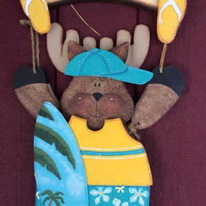 May include: Wooden moose figurine wearing a blue hat and yellow shirt holding a blue and yellow surfboard with a palm tree design. The moose is hanging from a wooden sign that says "Welcome".