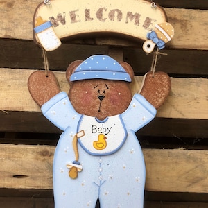 May include: Wooden welcome sign with a brown bear wearing a blue baby outfit with a yellow duck bib. The sign says "Welcome" and has a blue bottle and a rattle hanging from it.