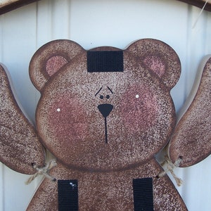 May include: A brown wooden bear with black eyes, a black nose, and a black stripe across its forehead. The bear is hanging from a string and has two brown wooden arms.