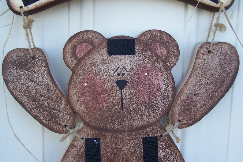 May include: A brown wooden bear with black eyes, a black nose, and a black stripe across its forehead. The bear is hanging from a string and has two brown wooden arms.