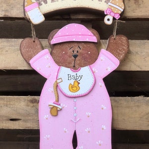 May include: A wooden welcome sign with the word "WELCOME" painted in brown letters. A pink teddy bear wearing a pink onesie with a yellow chick on the bib is holding a pacifier and a bottle. The sign is hanging from the bear's arms.