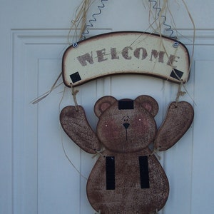 May include: A wooden bear holding a welcome sign. The bear is brown with black stripes and has a white face. The sign is cream colored with brown lettering that says "Welcome".