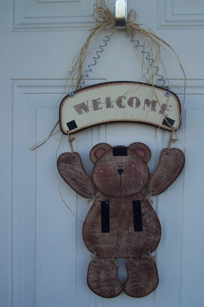 May include: A wooden bear holding a welcome sign. The bear is brown with black stripes and has a white face. The sign is cream colored with brown lettering that says "Welcome".