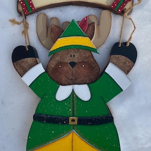 May include: Wooden Christmas door hanger featuring a green elf with a yellow hat and brown antlers. The elf is holding a sign that says "WELCOME" with red and green gift boxes on either side.