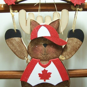 May include: A wooden moose wearing a red and white hat and shirt with a Canadian flag on the front. The moose is hanging from a wooden sign that says "WELCOME".