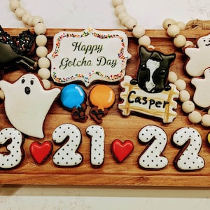 May include: A wooden tray holds decorated cookies, including a black horse, ghosts, balloons, and the text "Happy Gotcha Day." The cookies are arranged with the numbers "3 21 22" and hearts.