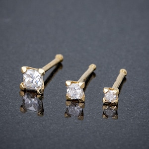 May include: Three gold nose rings with clear square-shaped gemstones. The rings are different sizes.