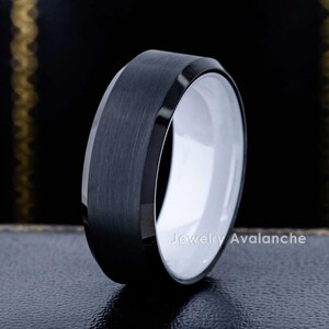 May include: A ring with a brushed matte black exterior and a smooth polished white interior. The band features beveled edges and a high-contrast design. The text Jewelry Avalanche is visible on the inner surface of the band, showcasing a modern aesthetic.