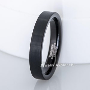 Puede incluir: Un anillo negro de carburo de tungsteno con acabado cepillado. El anillo tiene un perfil plano y un ancho de aproximadamente 3 mm. El texto "Tungsten Carbide" es visible en el interior del anillo. También es visible la marca "Jewelry Avalanche".