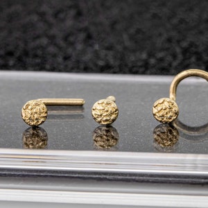 Puede incluir: Tres anillos de nariz de oro con un diseño de bola texturizada. Los anillos se muestran en una superficie negra.