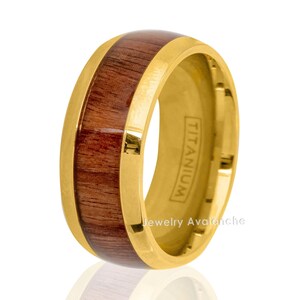 Puede incluir: Un anillo de titanio dorado con una incrustación de madera marrón. El anillo está grabado con la palabra "TITANIUM" y "Jewelry Avalanche".