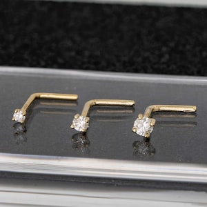 May include: Three gold nose rings with a small clear gemstone on each. The rings are on a black surface.