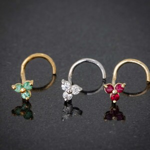 May include: Three gold nose rings with different colored gemstones. The first ring has three green gemstones, the second has three clear gemstones, and the third has three red gemstones.