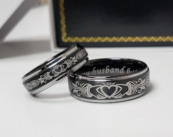 Könnte beinhalten: Zwei schwarze Ringe mit einem silberfarbenen Claddagh-Motiv, das Hände zeigt, die ein Herz halten, sowie filigrane Schnörkel. Die Innenseite des größeren Rings ist mit dem Text my husband 6-16 graviert und hat eine polierte Oberfläche.