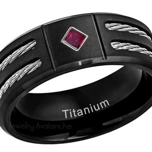 Op de afbeelding: Zwarte titanium ring met een rode edelsteen en een kabelontwerp. De ring is gegraveerd met het woord "Titanium" en de woorden "Jewelry Avalanche".