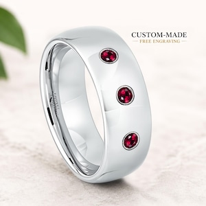 Könnte beinhalten: Ein polierter, silberfarbener Ring mit drei eingebetteten, runden, roten Edelsteinen. Der Ring hat ein glattes, abgerundetes Design und den Text "CUSTOM-MADE - FREE Engraving".