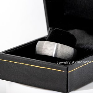 Peut inclure: Une bague argent&eacute;e avec une finition en m&eacute;tal bross&eacute; et une fine rayure blanche, pr&eacute;sent&eacute;e dans un &eacute;crin noir avec une bordure dor&eacute;e. La bague est au centre, mettant en valeur un design moderne.