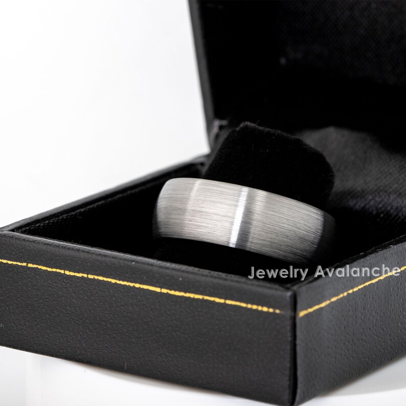 Peut inclure: Une bague argent&eacute;e avec une finition en m&eacute;tal bross&eacute; et une fine rayure blanche, pr&eacute;sent&eacute;e dans un &eacute;crin noir avec une bordure dor&eacute;e. La bague est au centre, mettant en valeur un design moderne.