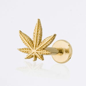 May include: A gold-colored, cannabis leaf-shaped earring stud. The stud features a detailed, textured leaf design and a round backing. The earring is shown against a white background, highlighting its metallic sheen.
