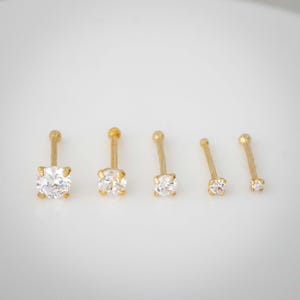 May include: Five gold nose rings with clear cubic zirconia stones. The rings are different sizes, with the largest ring on the left and the smallest ring on the right.