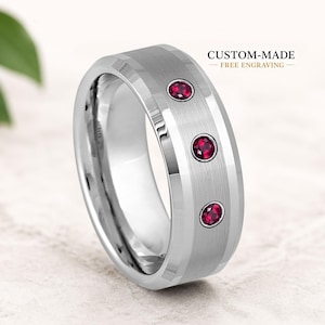 Könnte beinhalten: Ein silberfarbener Metallring mit gebürstetem Finish und drei runden, dunkelroten Edelsteinen, die vertikal entlang des Bandes gefasst sind. Der Text CUSTOM-MADE FREE ENGRAVING ist oben rechts im Bild über dem Ring zu sehen.