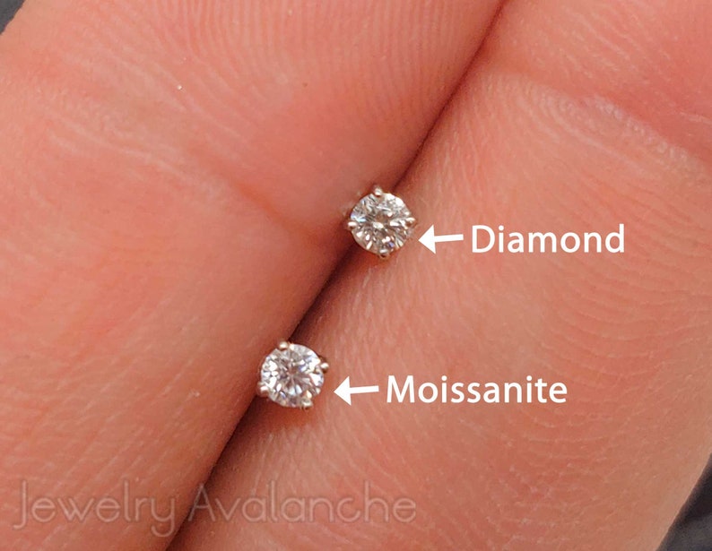 May include: Close-up of two small round gemstones set in metal. The gemstone on the left is labeled "Moissanite" and the gemstone on the right is labeled "Diamond".