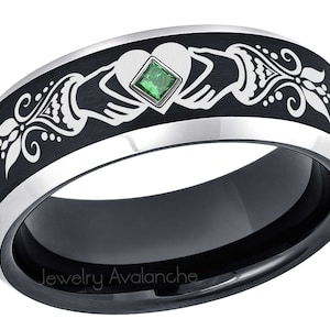 May include: A black and silver tungsten carbide ring with a Celtic knot design and a green emerald gemstone. The ring is engraved with the words "Jewelry Avalanche".