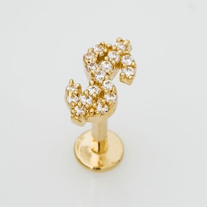 May include: A gold-colored lip piercing in the shape of a dollar sign, encrusted with clear, sparkling gemstones. The piercing has a flat, round base and a straight post.