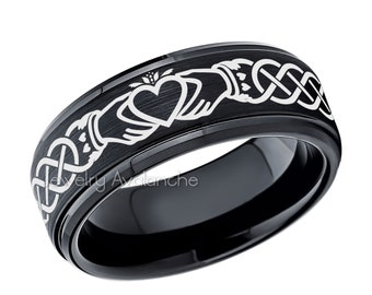 May include: A black ring with a white Claddagh design, featuring a heart, hands, and a crown. The ring has a polished, reflective surface and a brushed metal band with the Claddagh symbol. The ring is a symbol of love, loyalty, and friendship.