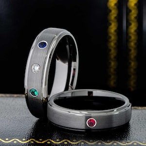 Op de afbeelding: Twee ringen van geborsteld zilverkleurig metaal met ingelegde ronde edelstenen. De ene ring heeft een blauwe, een doorzichtige en een groene steen, terwijl de andere ring een enkele rode steen bevat. Beide ringen hebben een gladde binnenkant.