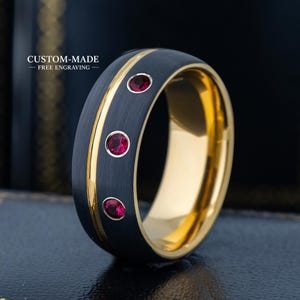 Puede incluir: Un anillo de metal cepillado negro con una ranura central dorada y tres piedras preciosas rojas redondas en línea vertical. El texto CUSTOM-MADE FREE ENGRAVING es visible en el lado izquierdo del anillo, que tiene un interior dorado pulido.
