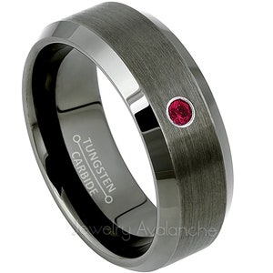 May include: A black tungsten carbide ring with a beveled edge and a single red gemstone set in the center. The ring is engraved with "TUNGSTEN CARBIDE".