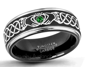 Könnte beinhalten: Ein schwarzer Ring aus Wolframkarbid mit einem silberfarbenen Claddagh-Design und einem zentralen grünen Edelstein. Das Band zeigt ein komplexes keltisches Knotenmuster um das Hände- und Herzmotiv, mit der Gravur TUNGSTEN CARBIDE im Inneren.