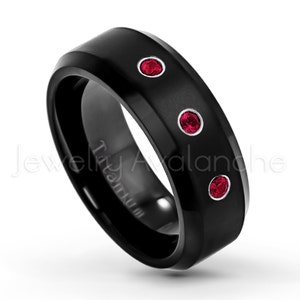 May include: A black titanium ring featuring a matte finish and three round, vibrant red gemstones set in silver-toned bezels along the outer band. The word Titanium is engraved on the inner surface of the band, which has a beveled edge design.