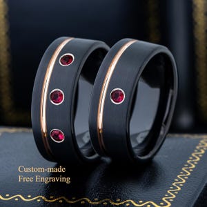 May include: Two black wedding bands with a rose gold stripe and three red gemstones. The rings are displayed on a dark surface. The text "Custom-made Free Engraving" is visible.